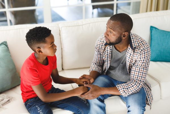 Serious african american father and son sitting on couch in living room talking and holding hands. family spending time at home, father son relationship.